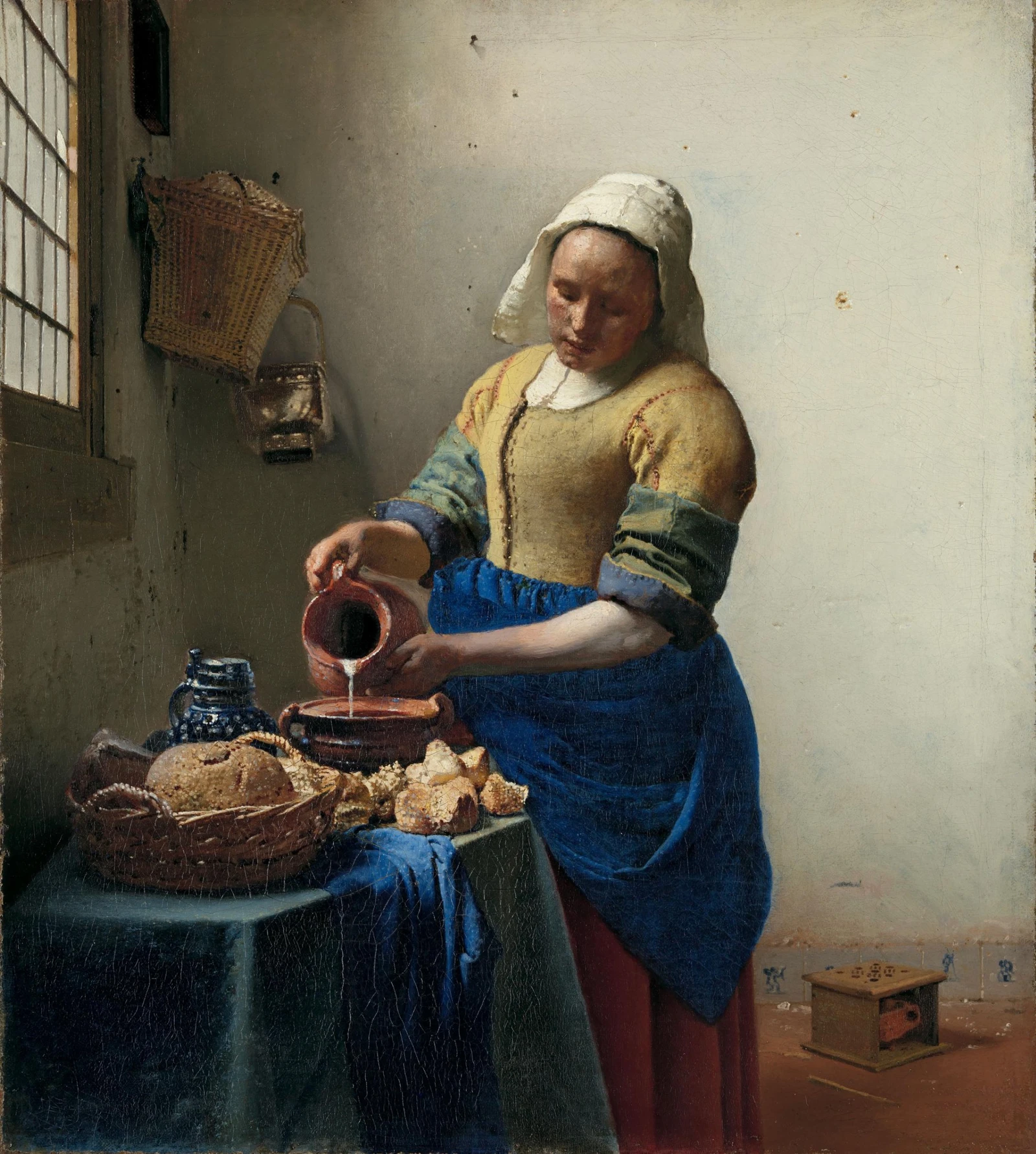 Uma mulher com touca branca, blusa amarela e avental azul despeja leite de uma jarra numa vasilha. À esquerda, uma janela ilumina a mesa com pães.