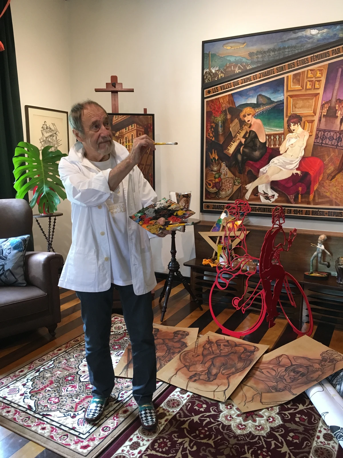 Juarez Machado com pincel e paleta, diante de uma pintura com duas mulheres e de esculturas vermelhas de ciclistas.